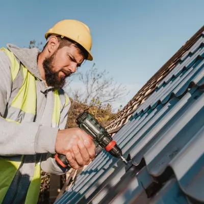 A construction worker in a yellow hard hat and reflective vest uses a power drill on a metal roof under a clear blue sky