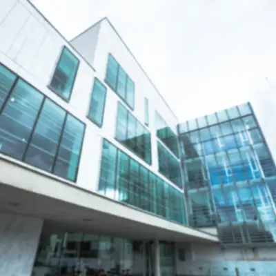 A modern, angular building with large glass windows reflecting a cloudy sky and surrounded by greenery